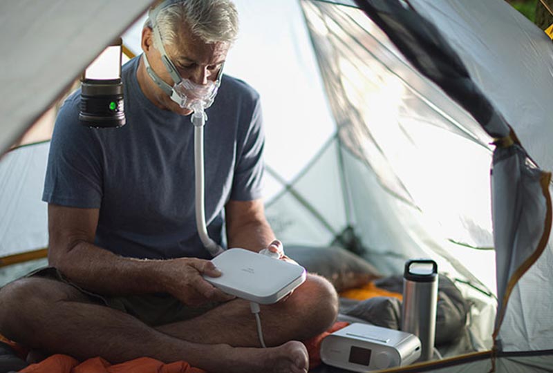 Mann sitzt mit der CPAP Maske auf in einem Zelt und stellt das CPAP Gerät an den externen Akku an