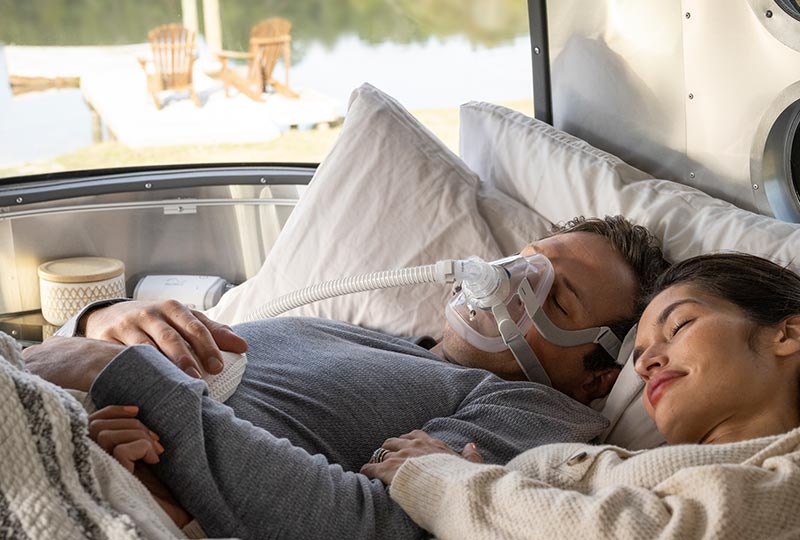 Pärchen liegt nebeneinander im Bett und schläft, der Mann hat eine CPAP Maske auf, das AirMini CPAP  Gerät liegt auf dem Nachttisch