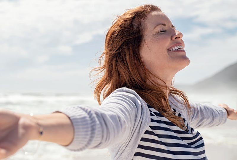 Frau  steht auf einem Strand, streckt beide Arme aus und lächelt dabei