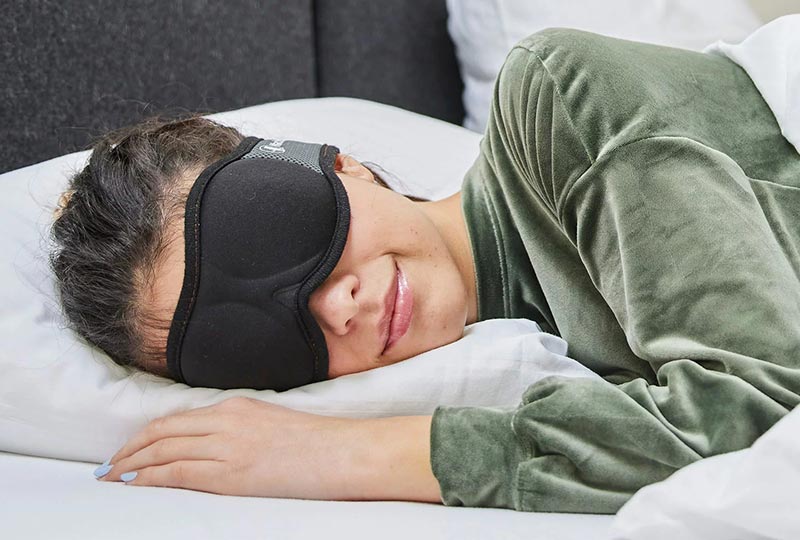 Dame liegt im Bett auf der Seite und schläft und hat dabei die ANINA Schlafmaske auf